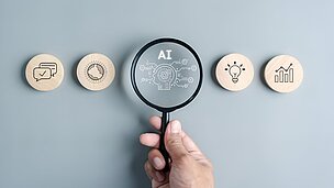 Symbolbild: Hand hält Lupe mit AI-Symbol über Icons für Kommunikation, Zeit, Ideen und Datenanalyse – Fokus auf Künstliche Intelligenz im Hotelkontext
