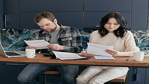 Zwei Menschen sitzen an einem Schreibtisch, wo zwei Kaffeebecher und ein Laptop stehen, und gucken sich beide ein eigenes Blattpapier an.