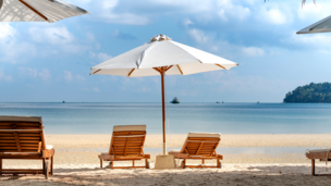 Man sieht einen Strand, wo ein Sonnenschirm und zwei liegen zu sehen sind