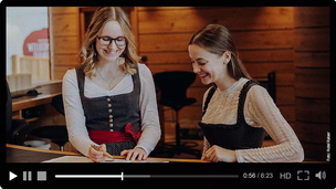 Zwei junge Frauen sitzen lächelnd an einem Holztisch und scheinen miteinander zu sprechen.