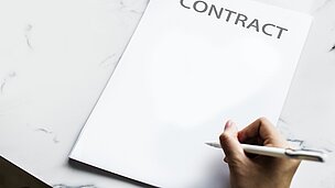 Auf dem Tisch liegt ein Vertrag. Über dem Vertrag sieht man, eine Hand die einen Stift hält