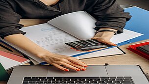 Jemand hat ein Taschenrechner in der Hand, unter dem Arm liegen Papiere und ein Laptop steht vor der Person auf dem Tisch