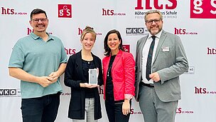 Vier Personen stehen nebeneinander vor einer weißen Fotowand mit roten und schwarzen Logos von „hts St. Pölten“ und „WKO“. Die zweite Person von links hält eine Smartphone-Box in den Händen und lächelt. Die beiden Frauen in der Mitte tragen schwarze beziehungsweise rote Kleidung; die Männer links und rechts sind in einem grünen Polo und einem grauen Anzug gekleidet. Die Atmosphäre wirkt freundlich und festlich, als würde ein Preis überreicht. Die Gesichter sind aus Datenschutzgründen nicht sichtbar.