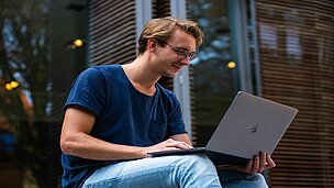 Ein Mann sitzt draußen vor einem Gebäude und hat sein Laptop auf dem Schos