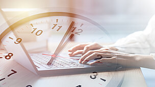Man sieht eine Person auf einen Laptop tippen und verschwommen sieht man auch eine Uhr 