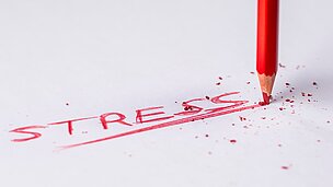 Ein roter Buntstift schreibt das Wort "Stress" auf ein weißes Blattpapier.
