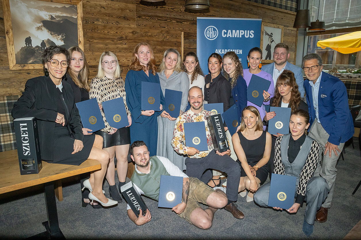 Auf dem Bild ist eine Gruppe von 16 Leuten zu sehen, die sich in einem Restaurant zusammengestellt haben, alle eine blaue Urkunde in der Hand halten und in die Kamera lächeln. Im Hintergrund ist ein blaues Rollup des ÖHV-Campus zu sehen.