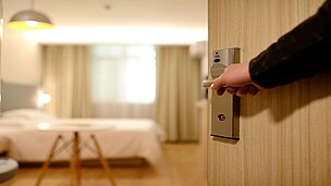 Man sieht wie eine Hand eine Tür aufmacht. Durch die Tür sieht man ein Hotelzimmer.