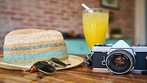 Man sieht auf einem Tisch liegen verschiedene Urlaubssachen wie: Ein Hut, eine Sonnenbrille, eine Kamera und ein Saft.